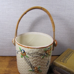 May include: Cream-colored ceramic basket with a woven texture and a brown rattan handle. The basket is decorated with floral designs in orange, blue, and green. The handle is attached with small, round, orange-brown accents.