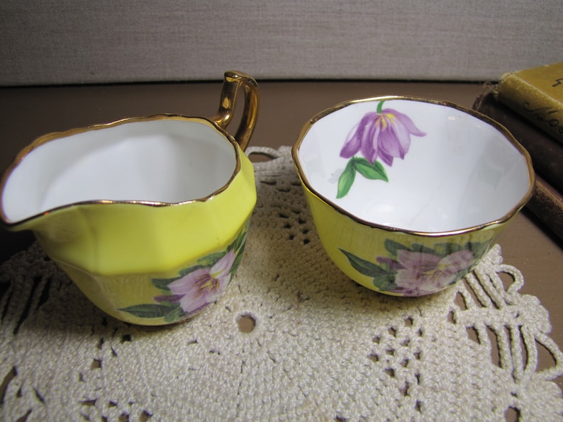 May include: A yellow creamer and sugar bowl set with a floral design. The creamer has a gold rim and a handle. The sugar bowl has a gold rim and a floral design on the side.
