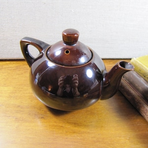 May include: A small brown ceramic teapot with a lid and a spout. The teapot is sitting on a wooden surface.