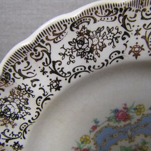 May include: A white ceramic plate with a gold and black floral design. The plate has a scalloped edge and a blue and gold center design.