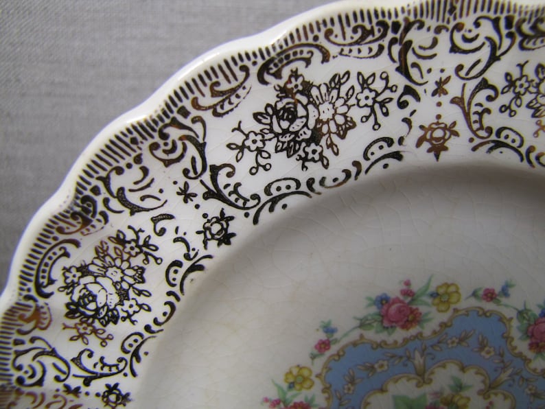 May include: A white ceramic plate with a gold and black floral design. The plate has a scalloped edge and a blue and gold center design.
