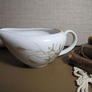 May include: White porcelain creamer with a wheat and floral design. The creamer has a curved spout and a handle. A gold trim accents the rim. The creamer is part of a vintage tableware set.
