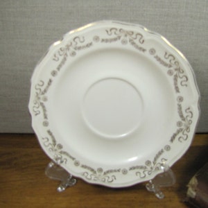 May include: White saucer with a gold rim and a delicate floral pattern. The saucer has a scalloped edge.