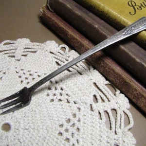 May include: A silver fork with a floral design on the handle. The fork is lying on a white crocheted doily.