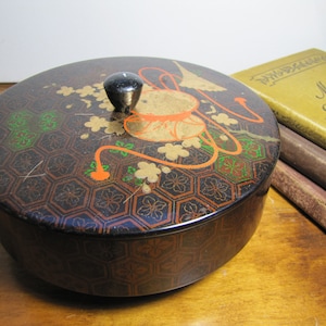 May include: A round, black lacquered box with a gold and orange floral design. The box has a black knob on the lid.