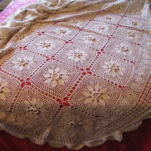 May include: A white crocheted tablecloth with a floral design, draped over a red bedspread.