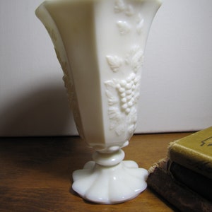 May include: A white milk glass vase with a raised design of grapes and leaves. The vase has a flared top and a pedestal base.