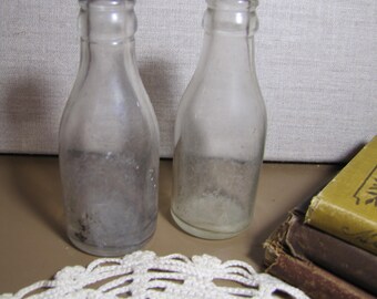 May include: Two clear glass bottles with a textured surface. The bottles are empty and have a vintage look.