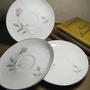 May include: Three white ceramic saucers with a delicate floral design featuring pink roses and gray leaves. The saucers have a silver rim and are stacked on top of each other.