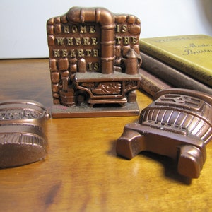 May include: Three small, copper-colored, decorative cast iron stove figurines. The center figurine has a brick wall behind it with the words "Home Where Hearth Is" written on it. The figurines are on a wooden surface.