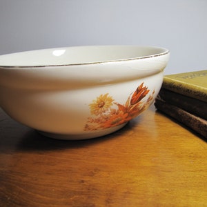 May include: A white ceramic mixing bowl with a gold rim and a floral design featuring orange and yellow flowers. The bowl is sitting on a wooden surface.
