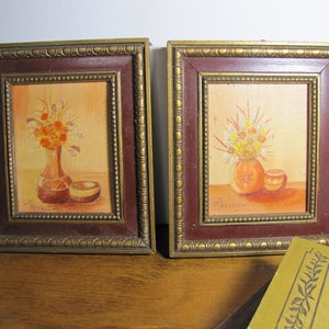 May include: Two framed paintings of floral arrangements in vases. The paintings are in a rustic style with a warm color palette. The frames are gold with a dark brown inner border.
