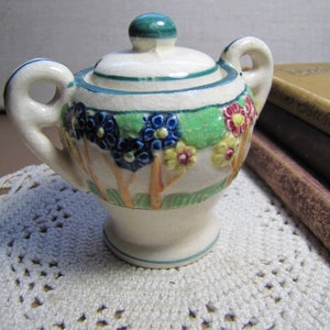 May include: A white ceramic sugar bowl with a green lid and floral design. The bowl has a floral pattern with blue, yellow, and red flowers. The bowl is sitting on a white crocheted doily.