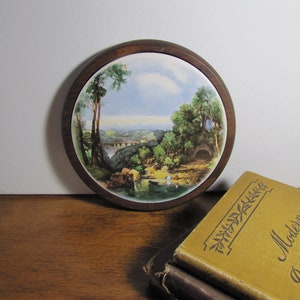 May include: A round wooden framed picture with a landscape scene of a river, trees, and a bridge. The picture is on a wooden surface.