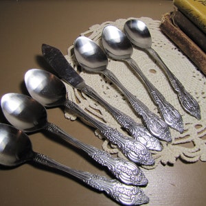 May include: A set of seven silver spoons and a butter knife with ornate handles. The spoons and knife are arranged on a white crocheted doily.