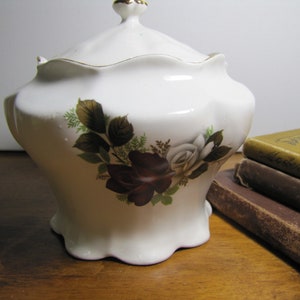May include: White ceramic jar with a lid, featuring a floral design of red and white roses with green leaves. The jar has a scalloped edge and a gold trim.