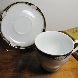May include: A white teacup and saucer with a black and gold rim and a seashell design. The saucer has a gold band around the edge.