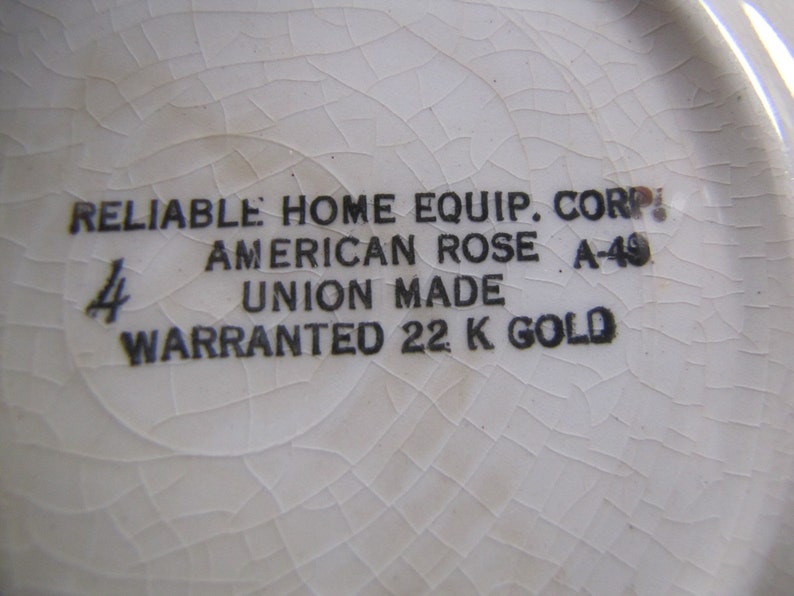 May include: Close-up of a white ceramic plate with black text that reads "RELIABLE HOME EQUIP. CORP. AMERICAN ROSE A-49 UNION MADE WARRANTED 22 K GOLD".