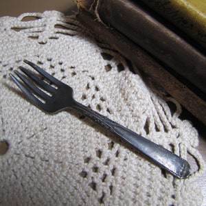 May include: A silver fork with a tarnished finish, lying on a white crocheted doily.