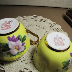 May include: Two yellow teacups with floral designs. The cups have gold trim and a red and white logo that reads "Royal Clarence China Made in England".