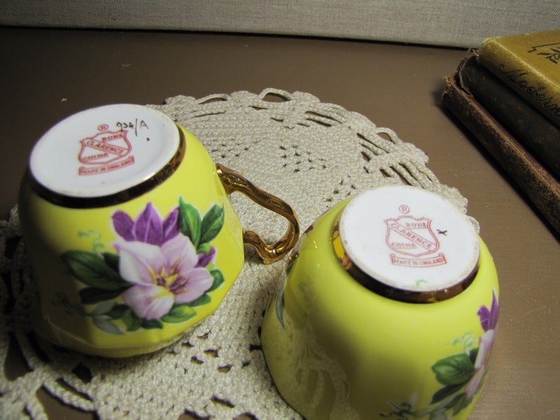 May include: Two yellow teacups with floral designs. The cups have gold trim and a red and white logo that reads "Royal Clarence China Made in England".