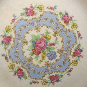 May include: A vintage ceramic plate with a delicate floral design. The center features a blue and gold floral motif with pink, yellow, and white flowers. The plate has a white background with a subtle textured finish.