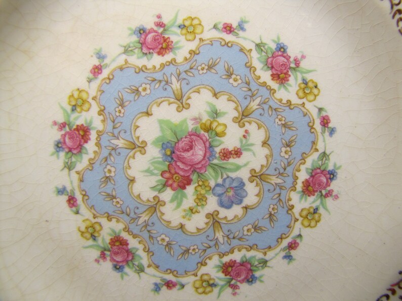 May include: A vintage ceramic plate with a delicate floral design. The center features a blue and gold floral motif with pink, yellow, and white flowers. The plate has a white background with a subtle textured finish.