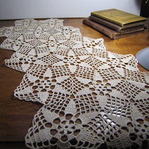 May include: A white crocheted table runner with a delicate floral pattern. The runner is laid out on a wooden table.
