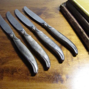 May include: Four silver butter knives with ornate handles. The knives are arranged in a row on a wooden surface.