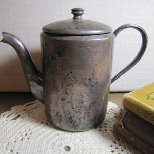 May include: A silver teapot with a lid and a handle. The teapot has a worn, aged look and is sitting on a white crocheted doily.