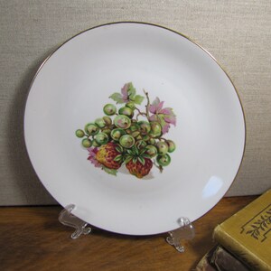 May include: White ceramic plate with a gold rim featuring a painted design of green grapes and red strawberries.
