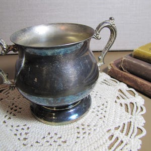May include: A silver sugar bowl with a rounded body and a single handle. The bowl is sitting on a white crocheted doily.