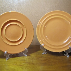 May include: Two orange ceramic plates on clear plastic stands. The plate on the left has a raised center circle. The plate on the right has a raised rim.