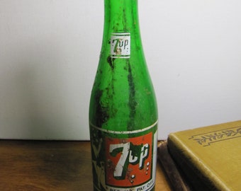 May include: A vintage green glass 7 Up bottle with a red and white label. The label says "7 Up" in white letters with a red background and "Reg. U.S. Pat. Off." in smaller white letters below the logo.