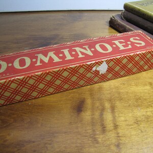 May include: A red and gold dominoes box with the words "DOMINOES" printed in gold letters. The box is made in the U.S.A.