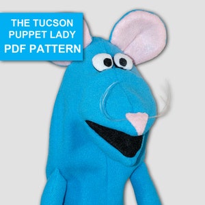 May include: A blue felt hand puppet shaped like a mouse with pink ears and a white moustache. The puppet has black eyes and a black mouth.
