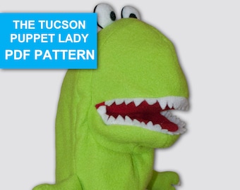 May include: Green felt hand puppet of a dinosaur with a large mouth and white teeth. The puppet has large white eyes and a green body. The text "THE TUCSON PUPPET LADY PDF PATTERN" is in a blue box at the top of the image.