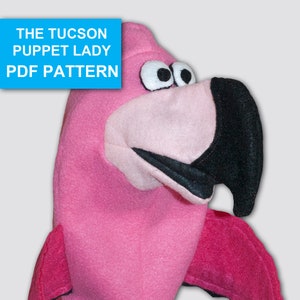 以下が含まれることがあります： 黒目の黒いクチバシを持つピンクのフラミンゴのパペット。パペットはピンクの体と翼を持っています。パペットはフェルト製で、パペットショーで使用できます。画像の上部には青いボックスに「THE TUCSON PUPPET LADY PDF PATTERN」というテキストがあります。