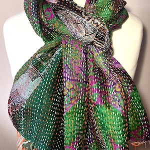 May include: A colourful, handmade scarf with a patchwork design. The scarf has a deep green base with purple, orange, and white stitching. The edges are finished with orange fringe. The scarf is draped over a mannequin.