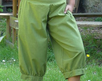 May include: A woman wearing a black crop top and green, wide-legged pants. She is standing in a grassy area with a wooden structure in the background.