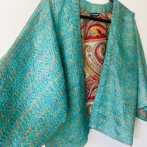 May include: A reversible open-front jacket featuring a teal exterior with a gold-toned intricate pattern and a contrasting interior lining in red, gold, and cream with a detailed paisley and bird motif. The garment has a label reading Poppa Mama.