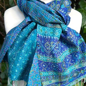 May include: A blue and teal scarf featuring intricate patterns and visible white running stitch embroidery throughout the fabric. The scarf has a soft, layered texture with frayed edges at the bottom, showcasing a mix of geometric and floral motifs.