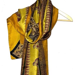 Peut inclure: Écharpe jaune moutarde ornée de motifs floraux et cachemire complexes en bordeaux, noir et marron. L'écharpe est drapée sur un cintre en bois, mettant en valeur son tissu fluide et son design détaillé. Les bords de l'écharpe sont bordés d'une rayure noire et bordeaux.