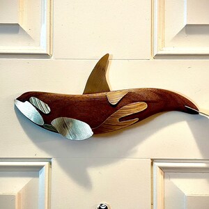 May include: A wooden orca sculpture with a dark brown body and silver accents. The orca has a dorsal fin and a tail fin. The sculpture is mounted on a white door.