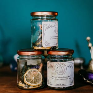 May include: Three clear glass jars with copper-coloured lids stacked in a pyramid. Each jar contains dried botanical ingredients, including visible dried lemon slices. The front-facing jars feature white rectangular labels with black text.