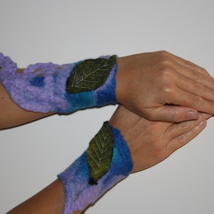 Puede incluir: Dos puños de lana fieltro con un degradado de color azul y morado. Cada puño presenta una aplicación de fieltro verde en forma de hoja.