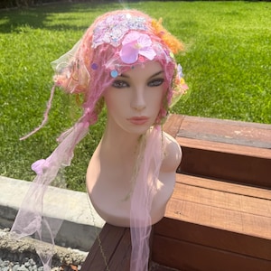 May include: A whimsical headpiece featuring a pink and orange color scheme. The headpiece is adorned with fabric flowers, sequins, and tulle. The design includes long, flowing strands and is displayed on a mannequin head.