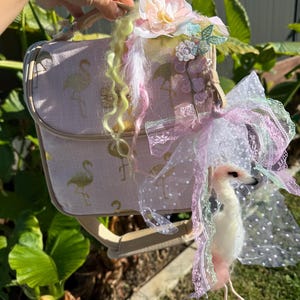 Op de afbeelding: Een roze handtas met een flamingo-patroon, versierd met bloemen en linten. De tas heeft een decoratieve flamingo-figuur en een schouderriem. De tas is versierd met roze, witte en gouden accenten.