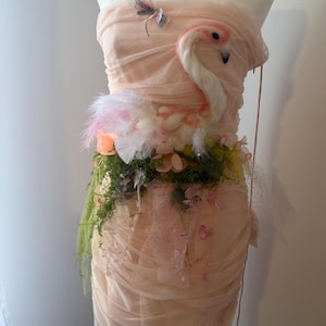 May include: A peach-coloured strapless dress on a mannequin, decorated with a flamingo, feathers, and floral details. A dragonfly is pinned to the bodice. The dress has draped fabric and a layered skirt, creating a whimsical, artistic design.