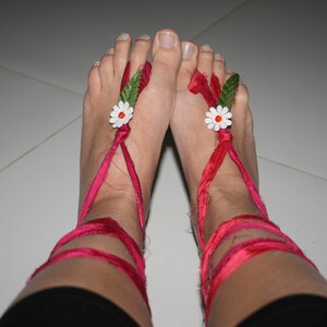 Può includere: Un paio di piedi che indossano sandali di nastro rosso con fiori bianchi e foglie verdi. I sandali sono legati alle gambe.